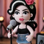 Crocheted doll with long black hair, a red flower accessory, wearing a black top and denim skirt, holding a microphone on a stand.