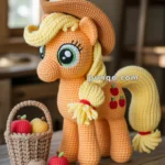 A crocheted pony with a yellow mane and tail, wearing a brown cowboy hat, is standing next to a woven basket filled with crocheted apples. The pony has big green eyes and small red apple patterns on its side, set indoors on a wooden table.