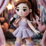 Crocheted doll with brown hair styled in a ponytail, wearing a light purple dress and boots, holding a microphone, standing in front of a backdrop with star and heart decorations.