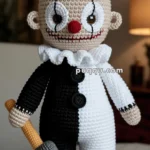 A crocheted clown doll with a half-black, half-white outfit, a black cutaway coat, and a small top hat, holding a plush hammer. The doll has a round red nose, large oval eyes, and a wide, stitched smile.