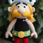 A handmade crocheted doll featuring a character with a winged helmet, large mustache, black shirt, red pants, and brown shoes.