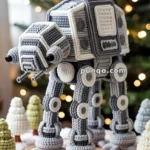 A crochet model of an AT-AT walker displayed in a winter-themed setting with small crocheted trees and snow, placed on a wooden surface with a blurred Christmas tree in the background with lights.