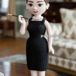 A crocheted doll dressed in a classic black dress with a pearl necklace and tiara, holding a long cigarette holder, standing on a wooden table in a room with elegant decor.