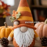 A crocheted gnome with a white beard and a tall orange and yellow hat, decorated with autumn leaves and sitting on a wooden table surrounded by pumpkins and pinecones.