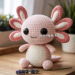 A crocheted pink axolotl toy with large black eyes and a smile, sitting on a wooden surface with a sprig of lavender in front of it. A plant and a curtain are in the background.