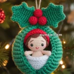 A crocheted Christmas ornament features a small doll with a red hat and a smiling face, nestled in a green wreath with holly leaves and red berries, hanging on a Christmas tree with lights.