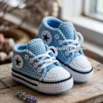 A pair of blue and white crocheted baby shoes designed to resemble sneakers, placed on a wooden surface near a window with a small purple flower nearby.