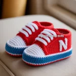 A pair of crocheted baby booties designed like sneakers, with red uppers, white laces, and blue soles, featuring a white letter 'N' on the side.