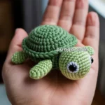 A crocheted green turtle with large eyes, resting in the palm of a hand.