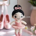 A hand holding a crochet keychain featuring a small ballerina doll with a pink tutu, black hair bun, and pink bow, set against a background with ballet shoes and pink flowers.