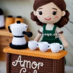 A crocheted doll standing behind a small, crafted table with the words 'Amor Cafe' written on it. The table holds a small crocheted coffee pot and cups. The doll has brown hair and is wearing a green outfit. Shelves with more crocheted cups are visible in the background.