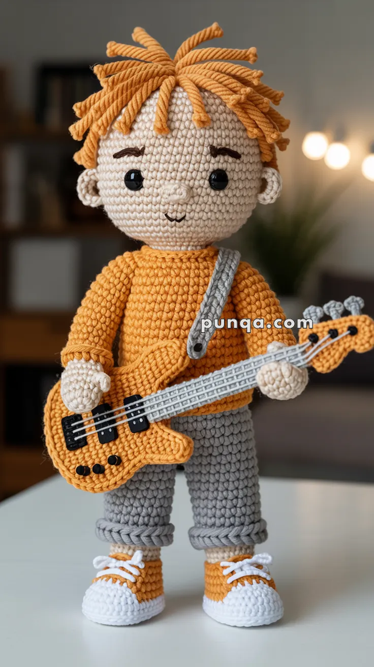 A crocheted doll with orange hair styled in spikes, dressed in an orange sweater and gray pants, holding a crocheted orange guitar with a gray strap. The doll is wearing orange and white sneakers and stands on a white surface with a blurred background.