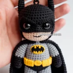 A hand holds a crochet keychain resembling a superhero with black ears, black and gray suit, and an emblem on the chest, featuring a yellow utility belt.