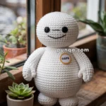 A crocheted stuffed doll resembling a rounded robot with black eyes and a smile, placed on a wooden windowsill next to potted succulents. The doll has a badge labeled "care" on its body.