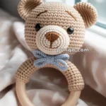 A crocheted teddy bear head on a wooden teething ring, adorned with a blue bow, resting on a soft patterned blanket by a window.