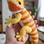 A hand holding a realistic crocheted bearded dragon in shades of yellow and orange, with intricate detailing on its scales and features, against a blurred indoor background.