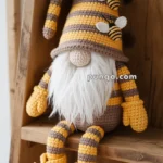 A crocheted gnome doll with a long, striped yellow and brown hat decorated with small bee designs, a white beard, and matching mittens and shoes, sitting on a wooden shelf.