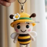 Hand holding a crocheted bee keychain with a yellow hat, white wings, and a flower decoration.