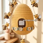 A crochet beehive adorned with small crocheted bees and flowers, labeled 'Sweet Honey,' hanging by strings in a sunlit room.