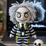Crocheted doll with wild gray hair wearing a black and white striped suit with green patches, next to a small crocheted skull on a wooden surface; a laptop with a blurred screen is in the background.