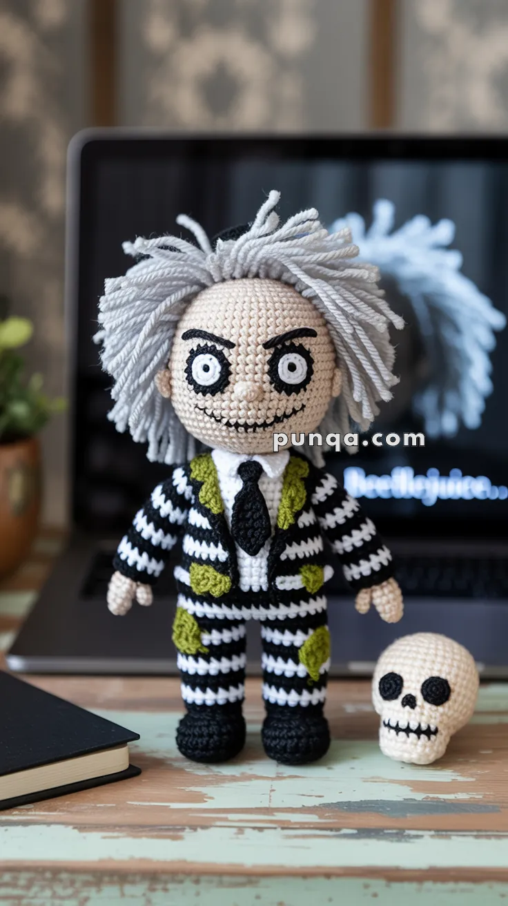 Crocheted doll with wild gray hair wearing a black and white striped suit with green patches, next to a small crocheted skull on a wooden surface; a laptop with a blurred screen is in the background.