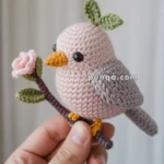 A hand holds a crocheted bird with light pink body, yellow beak and feet, grey wings, and black eyes, perched on a branch with green leaves and a pink flower.