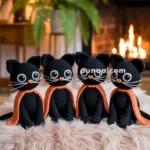 Four crocheted black cats with large eyes and orange bows sit on a fluffy stool in a cozy room with a fireplace and plants in the background.