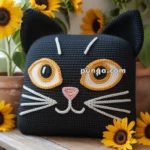 A knitted pillow designed to resemble a cat's face, with large orange and white embroidered eyes, sits on a wooden table surrounded by vibrant sunflowers in pots.
