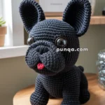Crocheted black dog plush sitting on a wooden stool with large eyes, a red tongue, and a sign in the background reading "Good Times and Frenchies."
