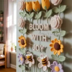 A handmade crochet wall hanging featuring flowers, bees, and the phrase 'Bloom with Joy.' The design includes yellow, pink, and purple flowers, green leaves, and dangling tassels, enhanced by the presence of small crochet bees.