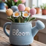 A crocheted blue watering can with the word "Bloom" stitched on it, filled with colorful crochet flowers, is placed on a wooden table with blurred flowers in the background.