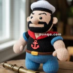 A crocheted sailor doll with a white sailor hat, black beard, and anchor emblem on its chest stands on a wooden table next to a brass telescope.