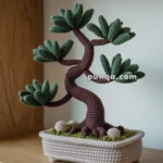 A crocheted bonsai tree with green leaves and a brown trunk, placed in a knitted planter on a wooden shelf.