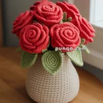 A handmade crochet arrangement featuring red roses and green leaves in a light-colored crocheted vase, placed on a wooden table near a window.