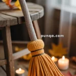 A hand-crocheted broom with a gray handle and an orange tassel rests against a wooden table, surrounded by scattered autumn leaves and lit tealight candles.