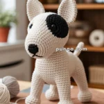 A crocheted dog toy with a black patch over one eye and a black nose, situated on a table with yarn and knitting needles nearby, in a cozy room setting.
