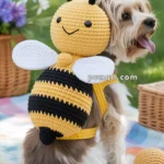 A small dog sits on a picnic blanket wearing a crocheted bee costume with wings and antennae in a park setting.