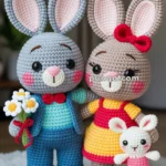 Two crocheted bunny dolls are standing on a wooden platform. The grey bunny, on the left, is wearing a blue suit and red bow tie, holding white and yellow flowers. The beige bunny, on the right, is dressed in a yellow and red dress with a red bow on its head, holding a smaller pink and white crocheted bunny.