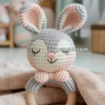 A soft, crocheted baby rattle shaped like a rabbit with closed eyes, featuring gray and pink yarn, attached to a wooden ring, resting on a fuzzy beige blanket with a nursery setting in the background.