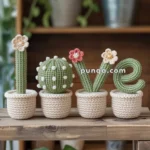 Four crocheted succulents in beige pots spell out "LOVE," each resembling a different cactus with small flowers, displayed on a wooden shelf with plants in the background.