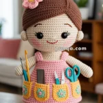 A handmade crochet doll with black eyes and a pink dress featuring pockets filled with sewing tools like scissors and a comb. The doll has brown hair styled in a bun adorned with colorful pins and a pink flower. It stands on a wooden surface in a cozy living room setting.