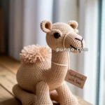 A crocheted camel plush toy sitting on a wooden surface near a window, with a small tag that reads "Handmade with love."