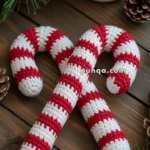 Two crocheted candy canes with red and white stripes on a wooden table, surrounded by pine branches, pinecones, and a mug with a festive design.