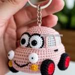 A hand holding a crocheted keychain shaped like a cute pink car with large, expressive eyes and colorful wheels.
