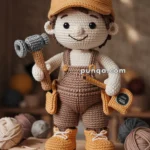 A crochet carpenter doll resembling a construction worker, wearing a brown cap, overalls, and yellow boots, holding a hammer and a measuring tape, standing on a wooden table labeled 'Handmade with Love.'
