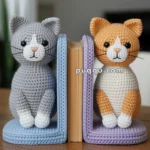 Two crocheted cat bookends, one gray and one orange, positioned on either side of an upright book, against a blurred indoor background.
