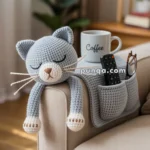 A crocheted gray cat with closed eyes is draped over the arm of a beige sofa, next to a mug labeled "Coffee" and an armrest pocket holding a remote control and glasses.