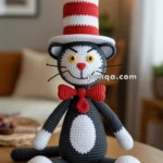 A crocheted toy cat dressed in a red and white striped hat and a red bow tie, sitting on a wooden table in a warmly lit room.