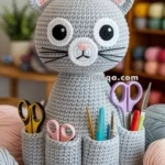 A crocheted grey cat-shaped organizer with large eyes and pink ears, holding various craft tools like scissors and knitting needles in its pockets, set on a workspace filled with yarns and crafting materials.