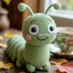 A crocheted green caterpillar toy with large eyes and a smiling face, sitting on a wooden surface surrounded by autumn leaves and a floral teacup in the background.